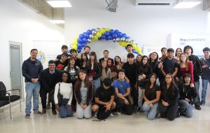 "Grupo de estudiantes y docentes del Preuniversitario UC posando juntos en una sala, bajo un arco de globos azules, amarillos y plateados.
